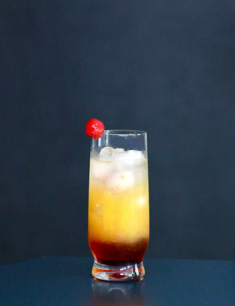 Italian Sunset Cocktail in einem hohen Glas mit Eis, Farbverlauf von Orange zu Rot, garniert mit einer Cocktailkirsche am Glasrand.