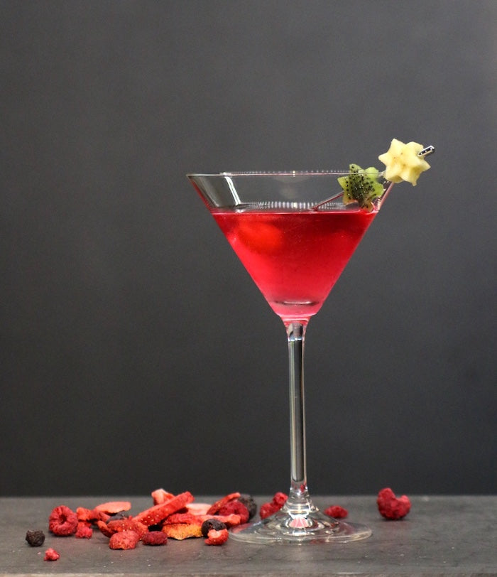 Huapala Cocktail in einem Cocktailglas, leuchtend rot, garniert mit Kiwi- und Sternfruchtspieß, umgeben von getrockneten Beeren.