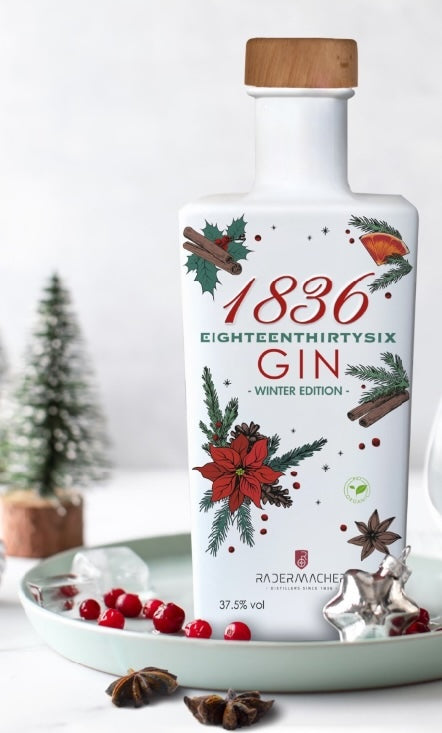 1836 Gin Winteredition in weißer rechteckiger Flasche auf winterlicher Dekoration mit Gewürzen, Cranberries und kleinen Tannen.