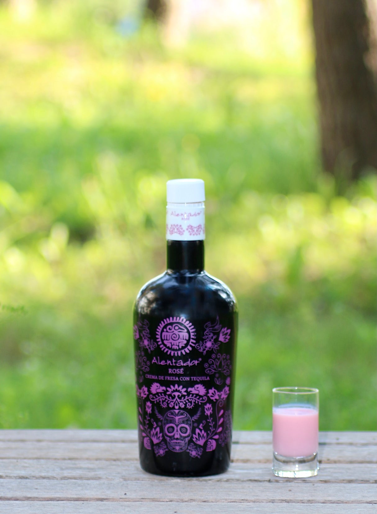 Schwarze Flasche Alentador Rosé Crema de Fresa con Tequila Likör mit lila Motiv, daneben gefülltes Shotglas auf Holztisch.