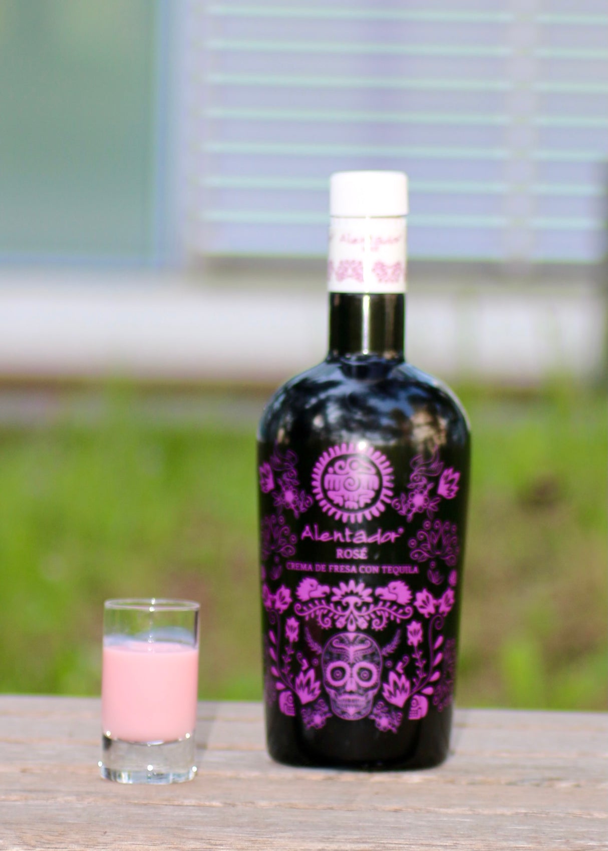 Schwarze Flasche Alentador Rosé Crema de Fresa con Tequila Likör mit lila Design, daneben gefülltes Shotglas auf Holztisch