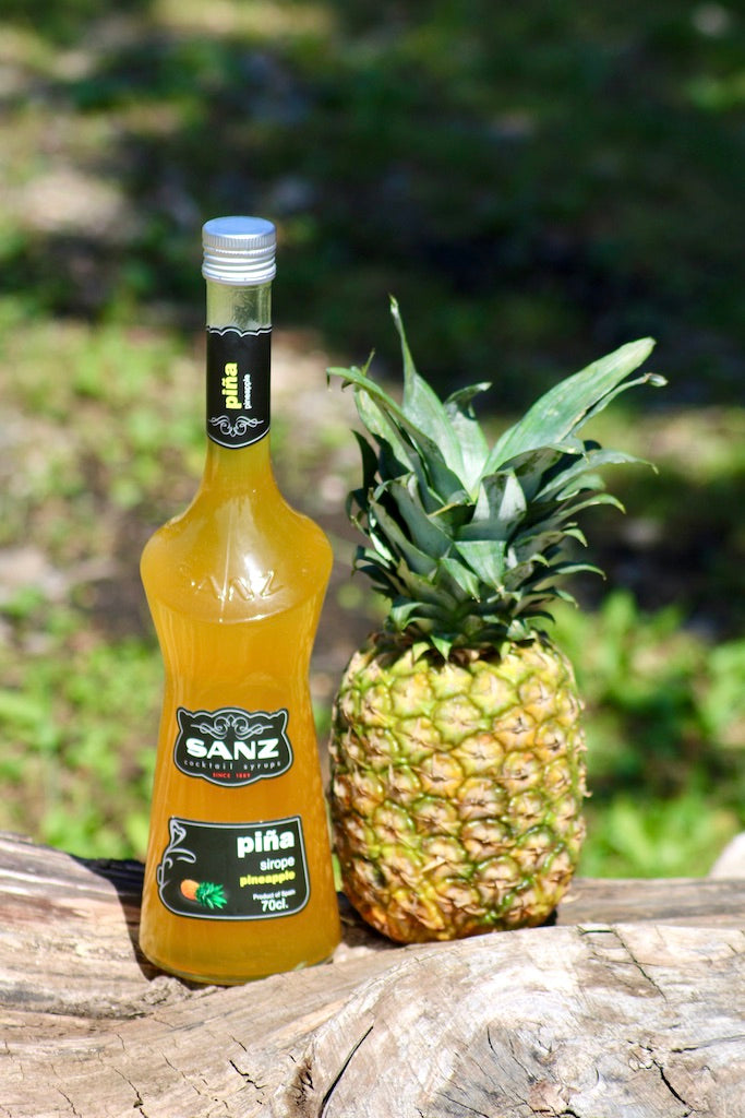Ananassirup Sanz 0,7L neben einer frischen Ananas auf einem Holzstamm, mit natürlichem Hintergrund.