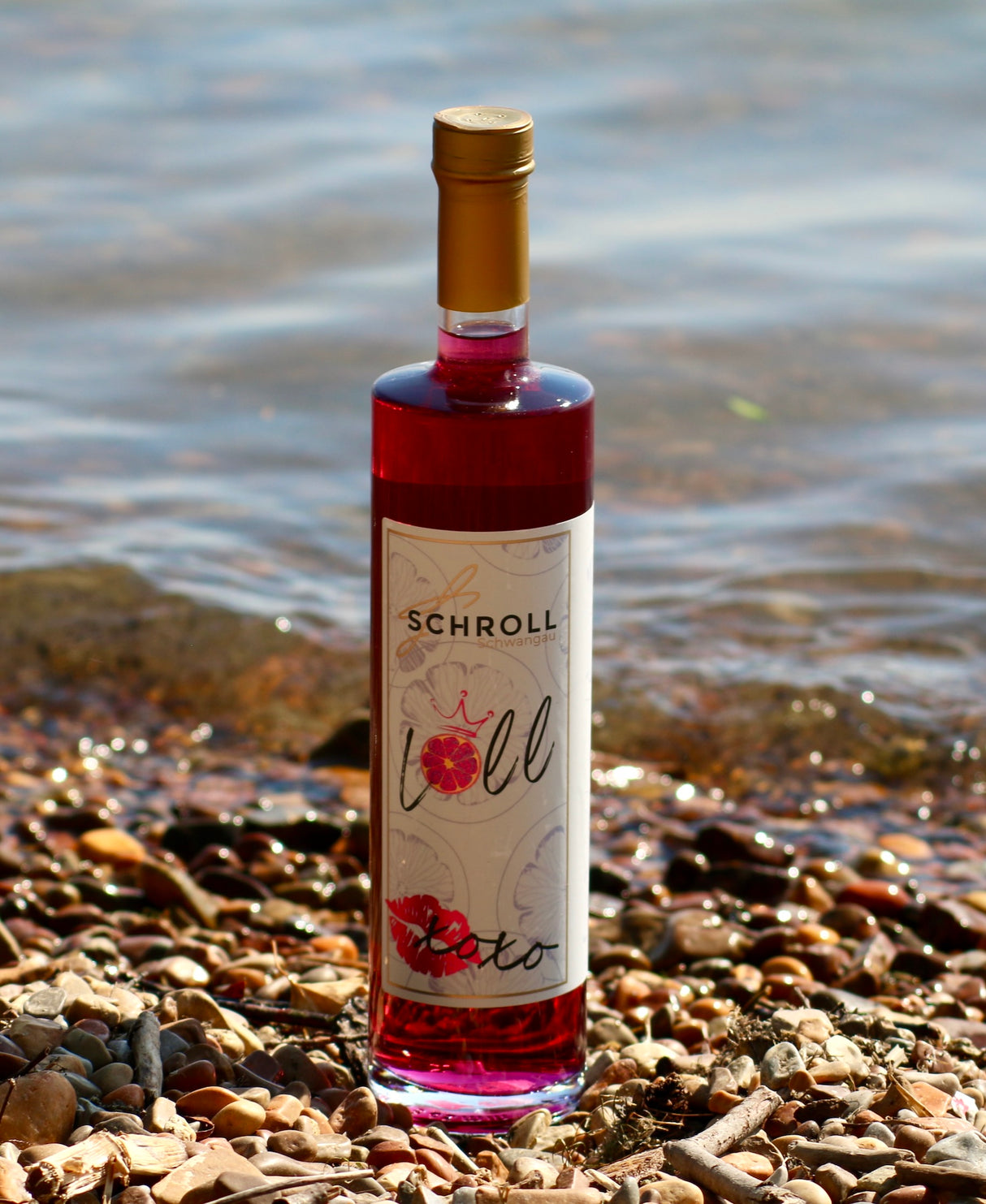 Flasche Loll Aperitif von Schroll am Ufer mit Kieselsteinen und Wasser im Hintergrund, bei Sonnenlicht aufgenommen.