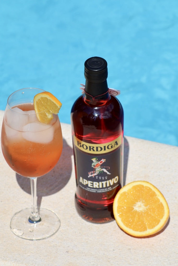 Aperitivo Spritz im Weinglas mit Eis und Orangenscheibe neben Bordiga Aperitivo Flasche und halber Orange am Poolrand.