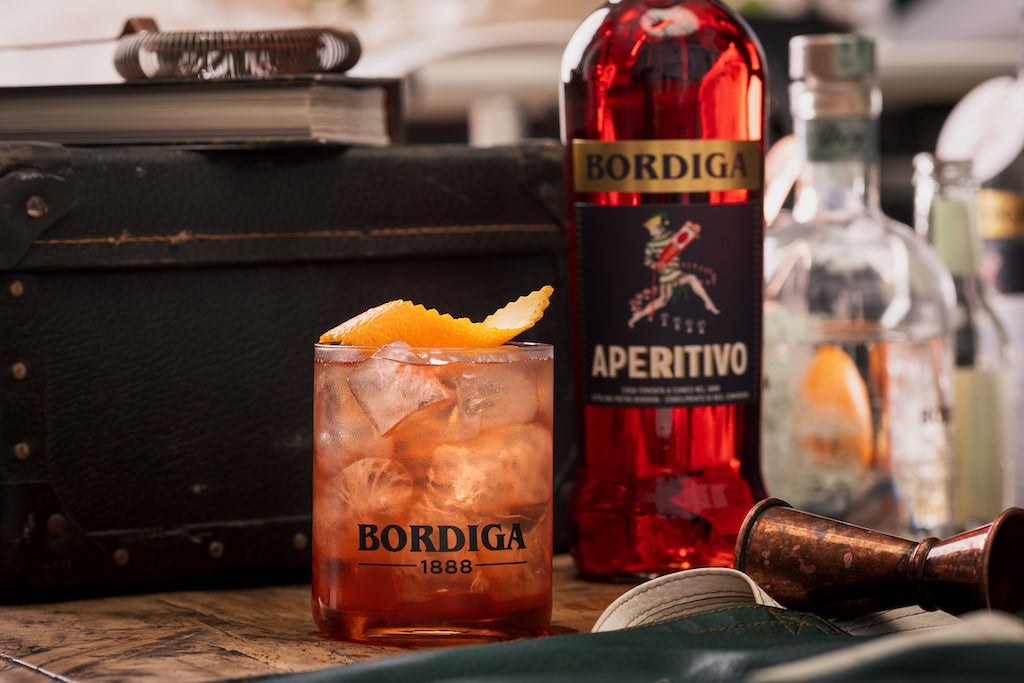 Cocktailglas mit Eis und Orangenzeste, daneben eine Flasche Bordiga Aperitivo, im Hintergrund Barzubehör und Flaschen.