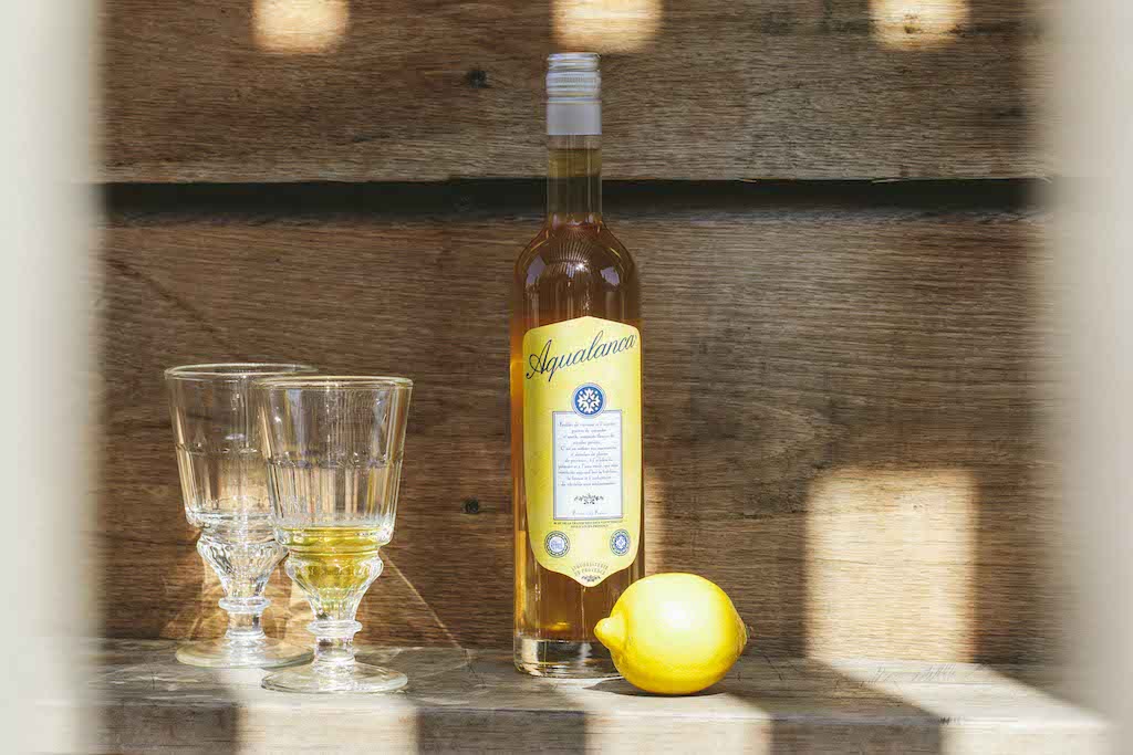 Flasche Aqualanca L'Anis d'Antan mit zwei Gläsern und einer Zitrone auf einem Holztisch im Sonnenlicht