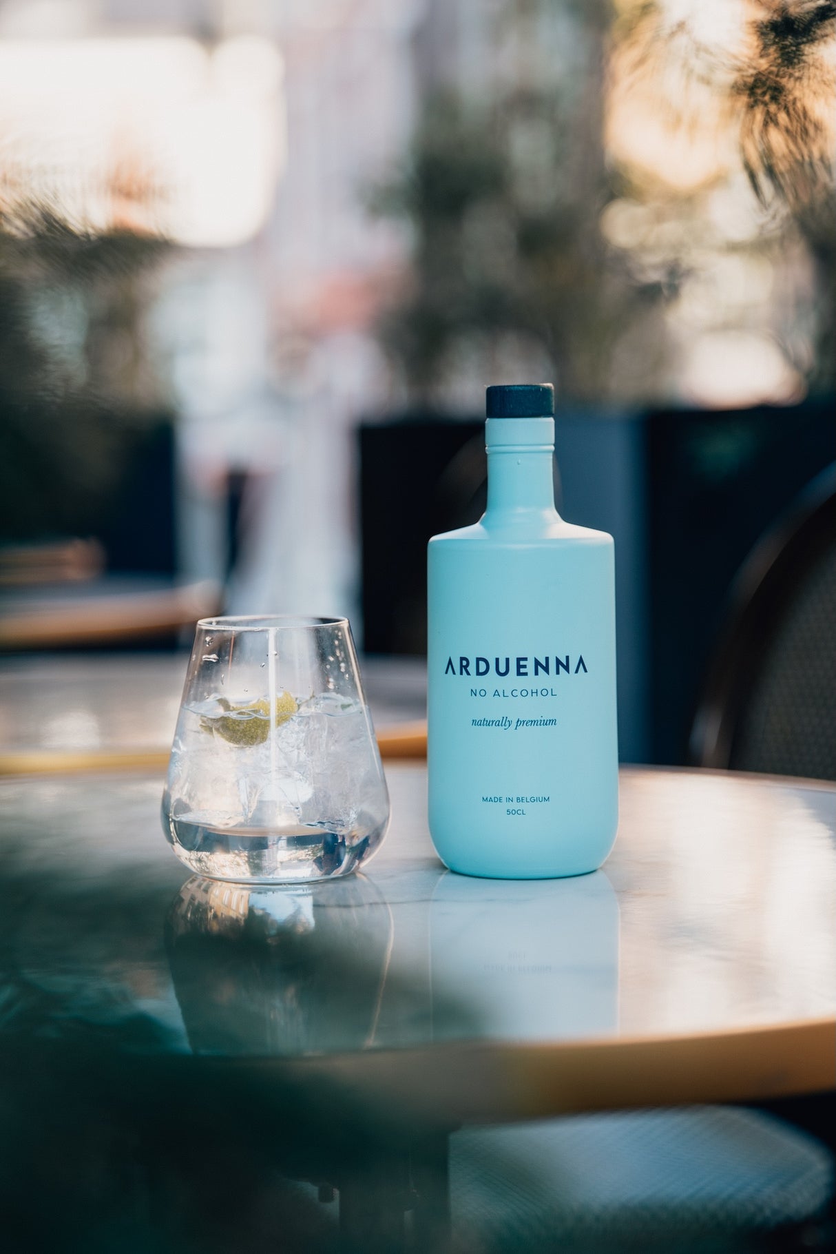 Arduenna No Alcohol 0,5 l neben einem Glas mit Tonic, Eis und Limette auf einem runden Tisch mit unscharfem Hintergrund