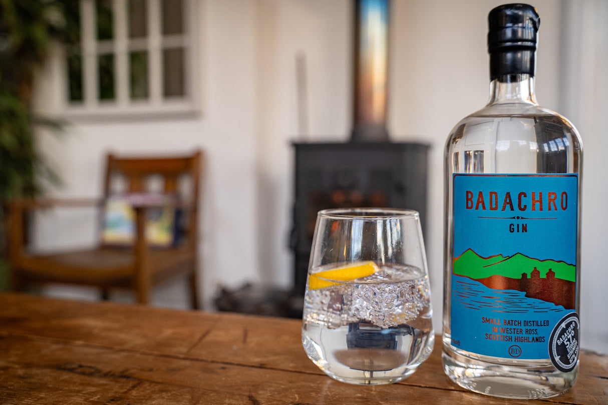 Flasche Badachro Storm Strength Gin neben einem Glas Gin & Tonic mit Zitronenzeste auf einem Holztisch, im Hintergrund ein Kamin.