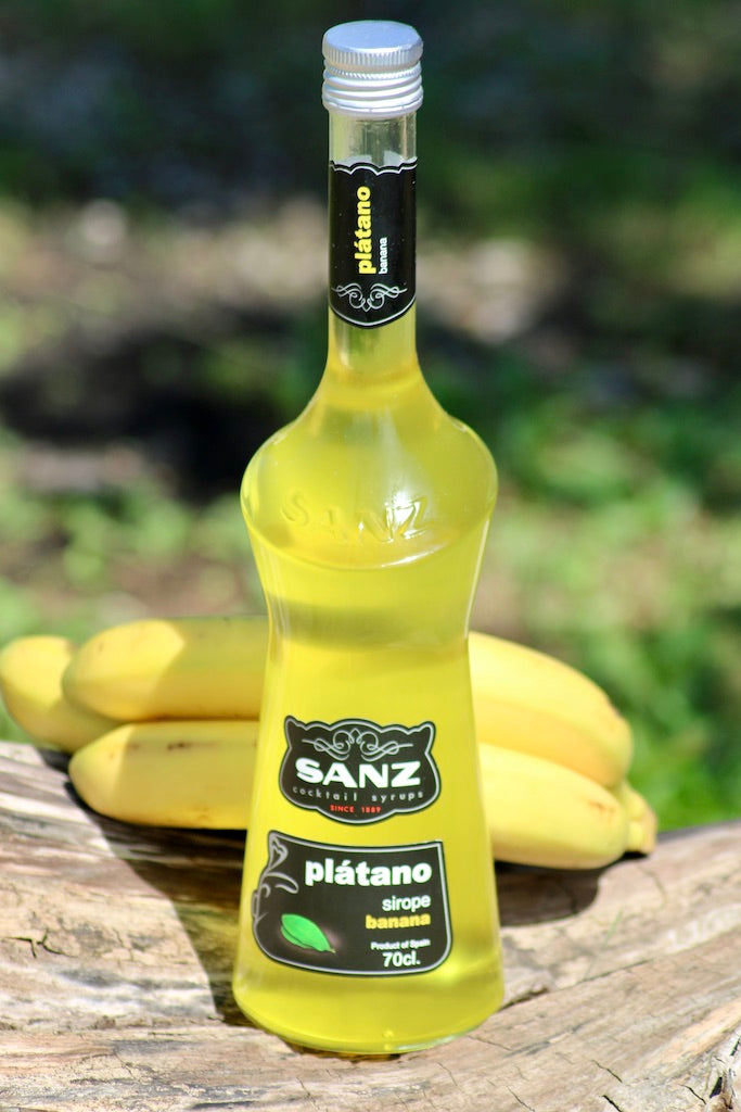 Flasche Sanz Plátano (Banane) Sirup, gelber Sirup in 700 ml Flasche, vor frischen Bananen auf einem Baumstamm