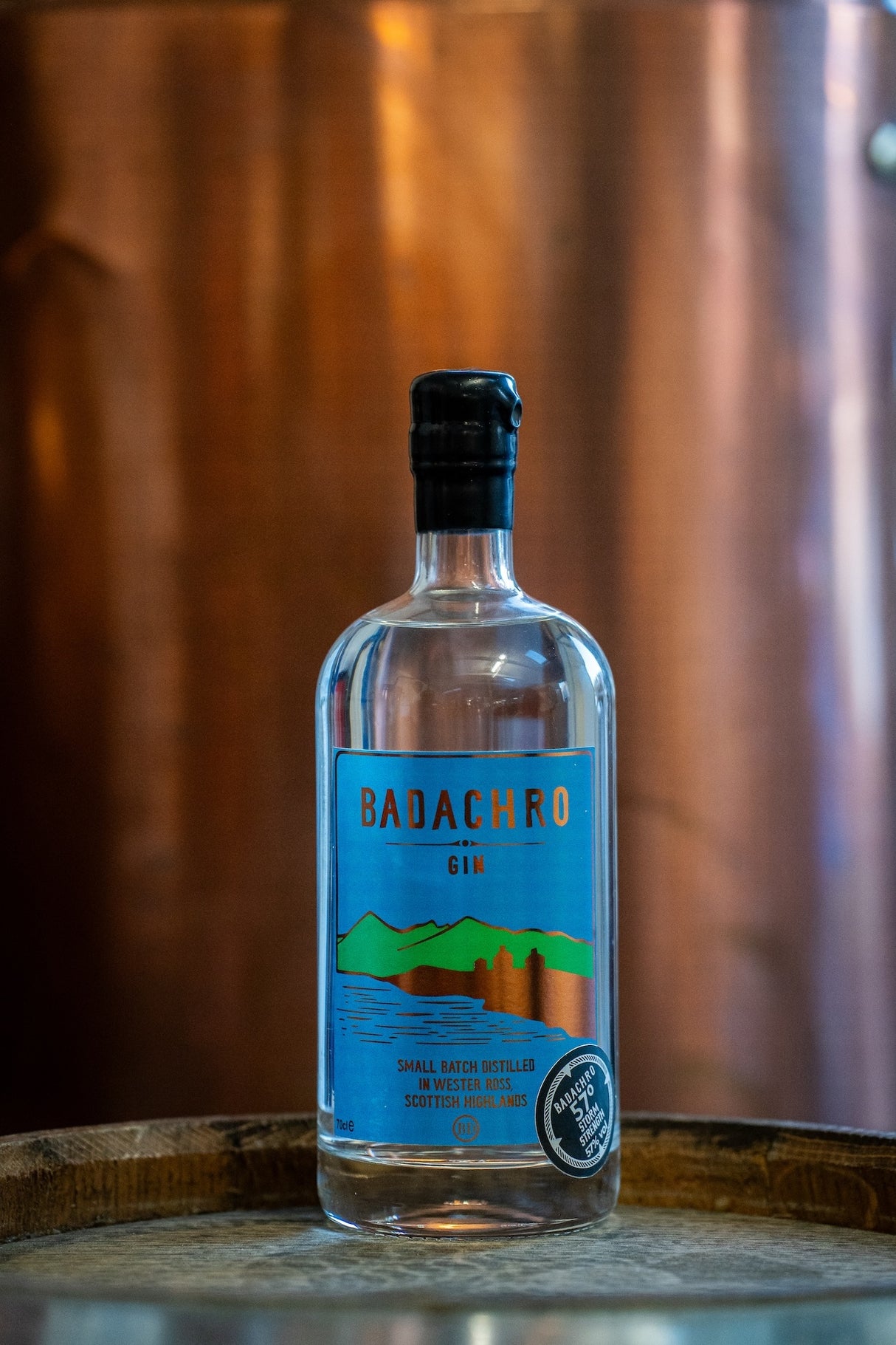 Flasche Badachro Storm Strength Gin auf einem Holzfass vor einem Kupfer-Destillationsapparat in der Destillerie.