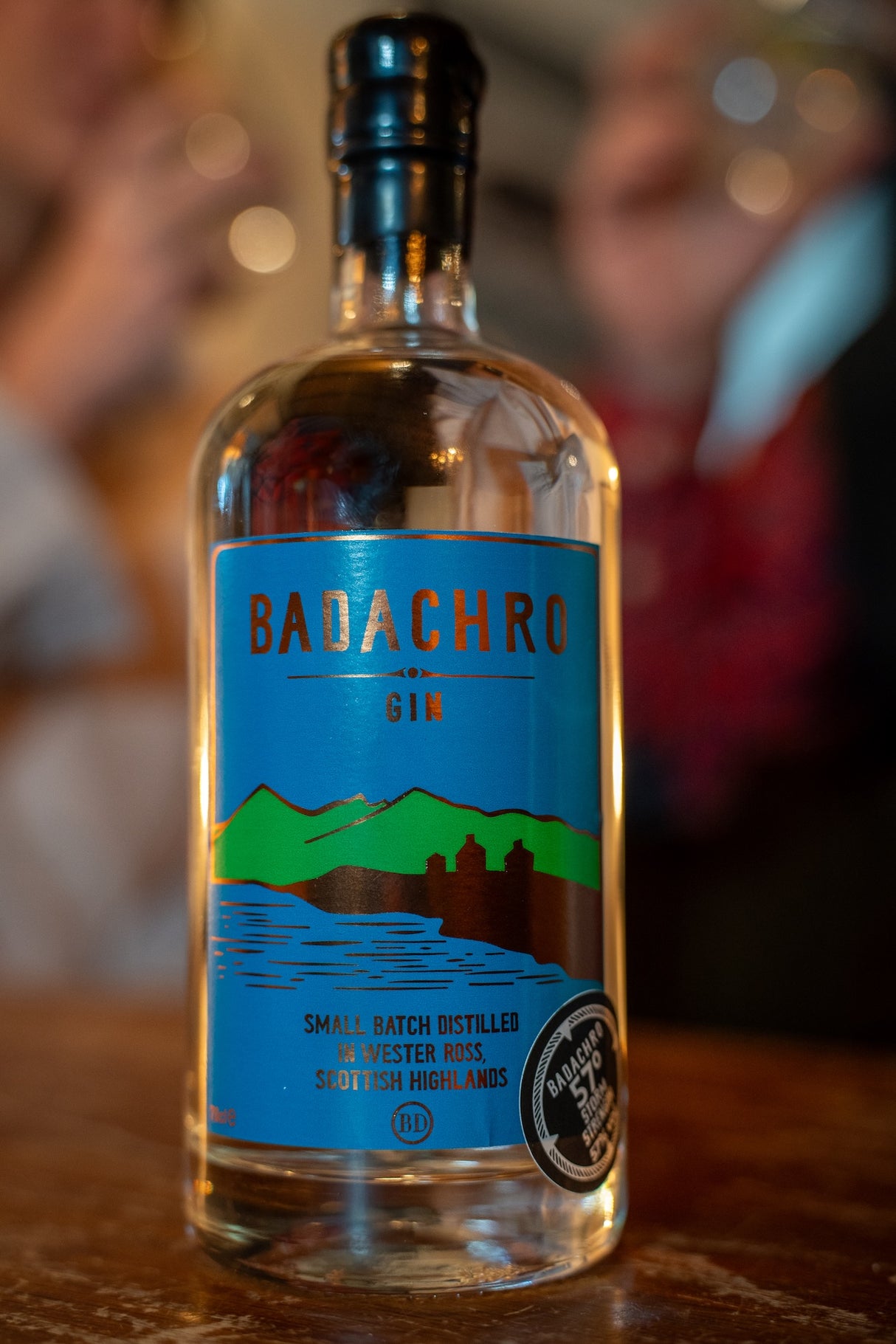 Flasche Badachro Storm Strength Gin auf einem Holztisch, im Hintergrund unscharf Personen mit Gläsern.