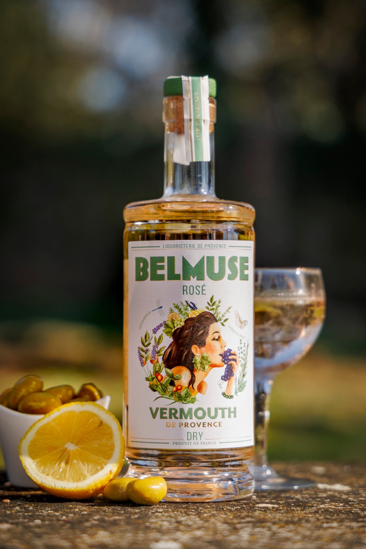 Belmuse Rosé Wermut aus der Provence, Flasche vor Glas, Zitrone und Oliven auf Steinuntergrund bei Tageslicht