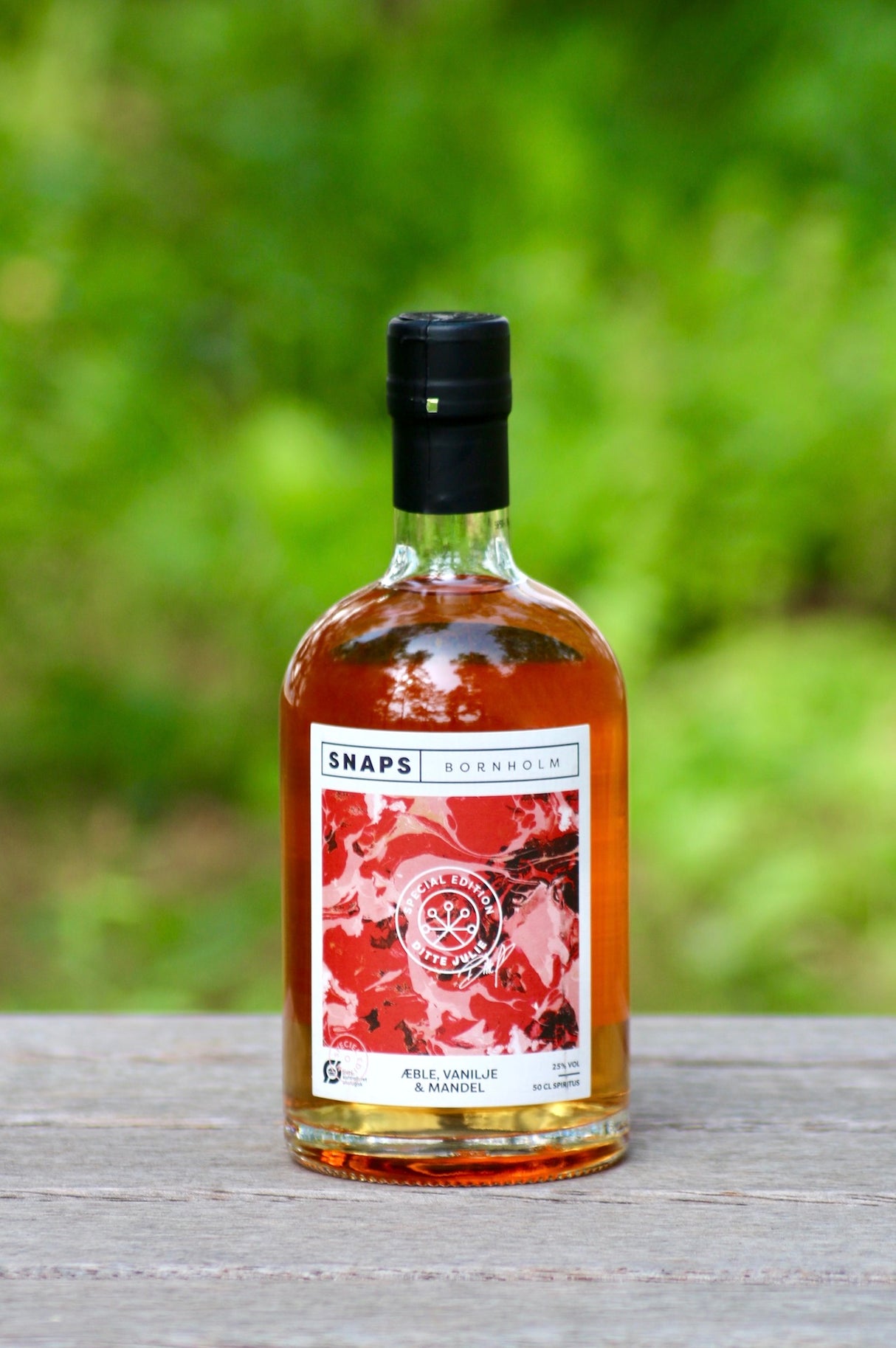 Eine Flasche Bio-Schnaps von Bornholm, mit den Geschmacksnoten Apfel, Vanille und Mandel, steht auf einer Holzoberfläche vor einem unscharfen, grünen Hintergrund.