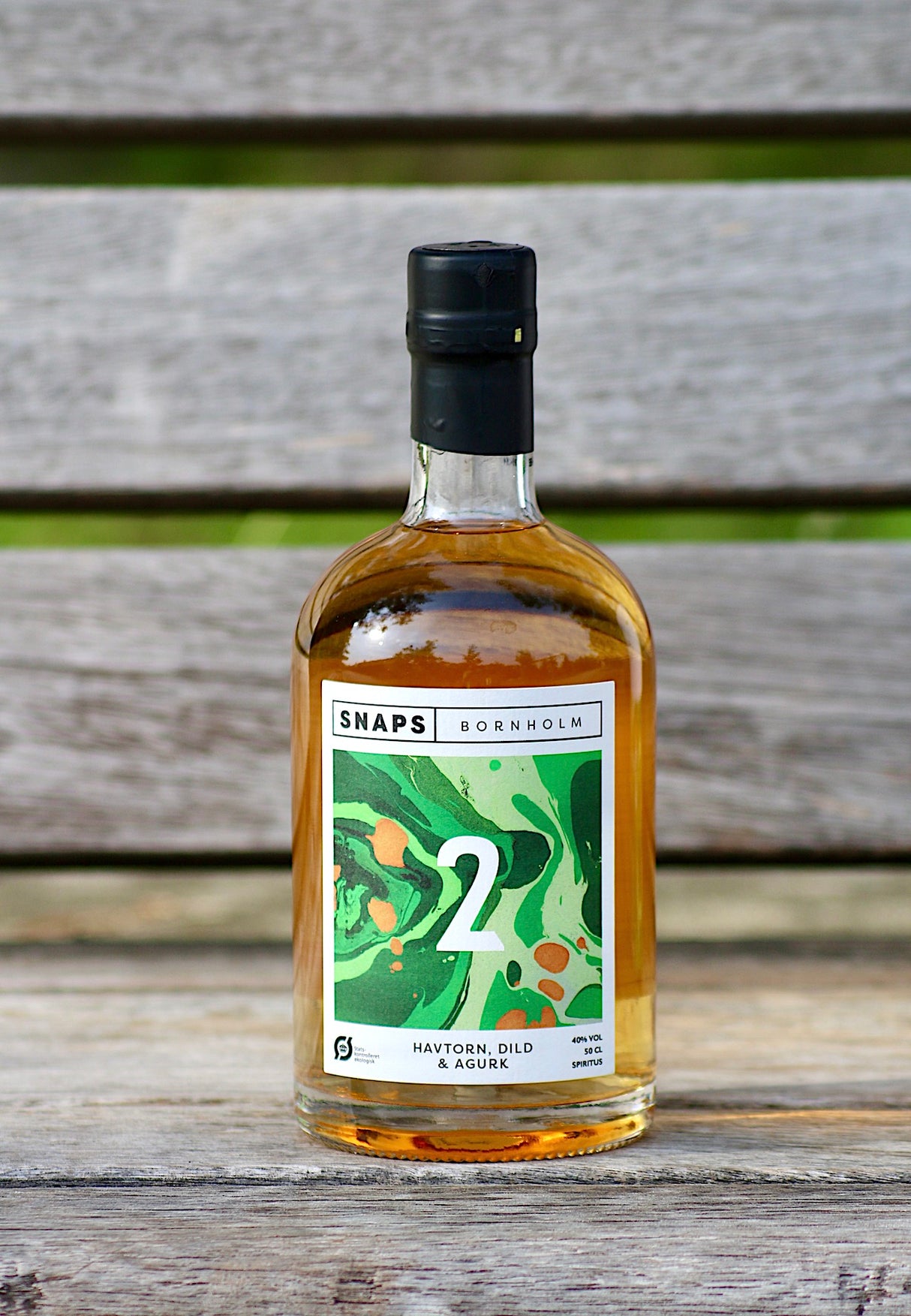 Eine Flasche Bio-Schnaps von Bornholm mit den Geschmacksnoten Sanddorn, Dill und Gurke, stehend auf einer Holzbank vor einem neutralen Hintergrund.