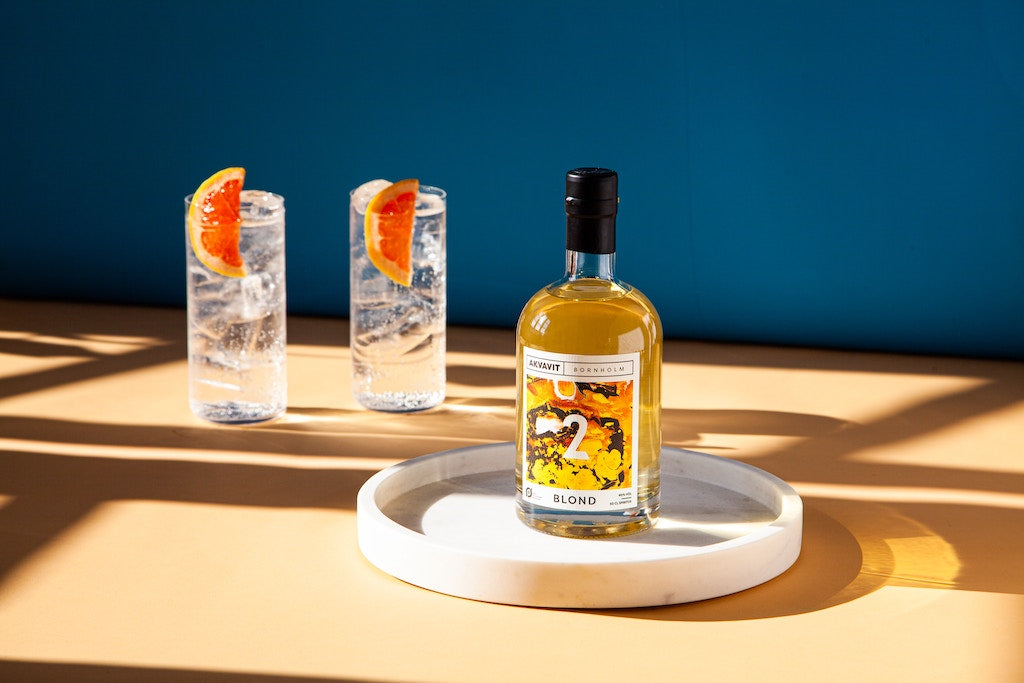 Bornholm Blond Akvavit auf weißer Platte, davor zwei Longdrinks mit Eis und Grapefruitscheiben auf hellem Tisch.