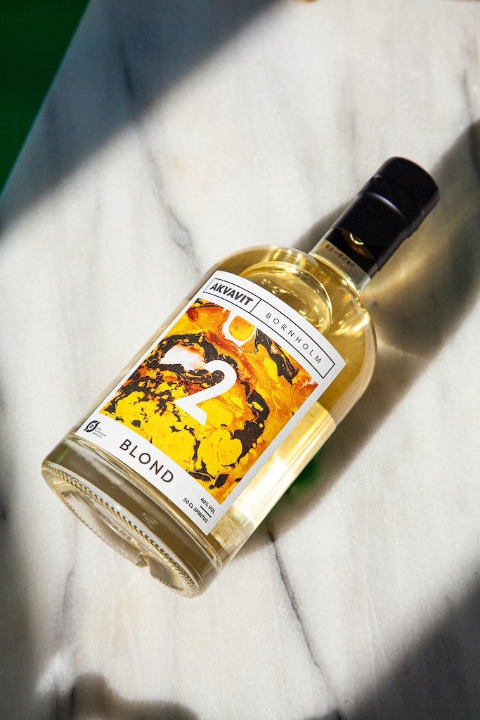 Bornholm Blond Akvavit-Flasche mit gelb-orangefarbenem Etikett, liegend auf hellem Marmorboden mit Lichteinfall.