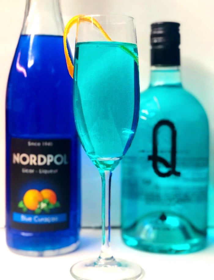 Blue Gin Fizz Cocktail mit blauer Curacao-Likörflasche, Ginflasche und Orangenzeste im Sektglas, leuchtend blau.