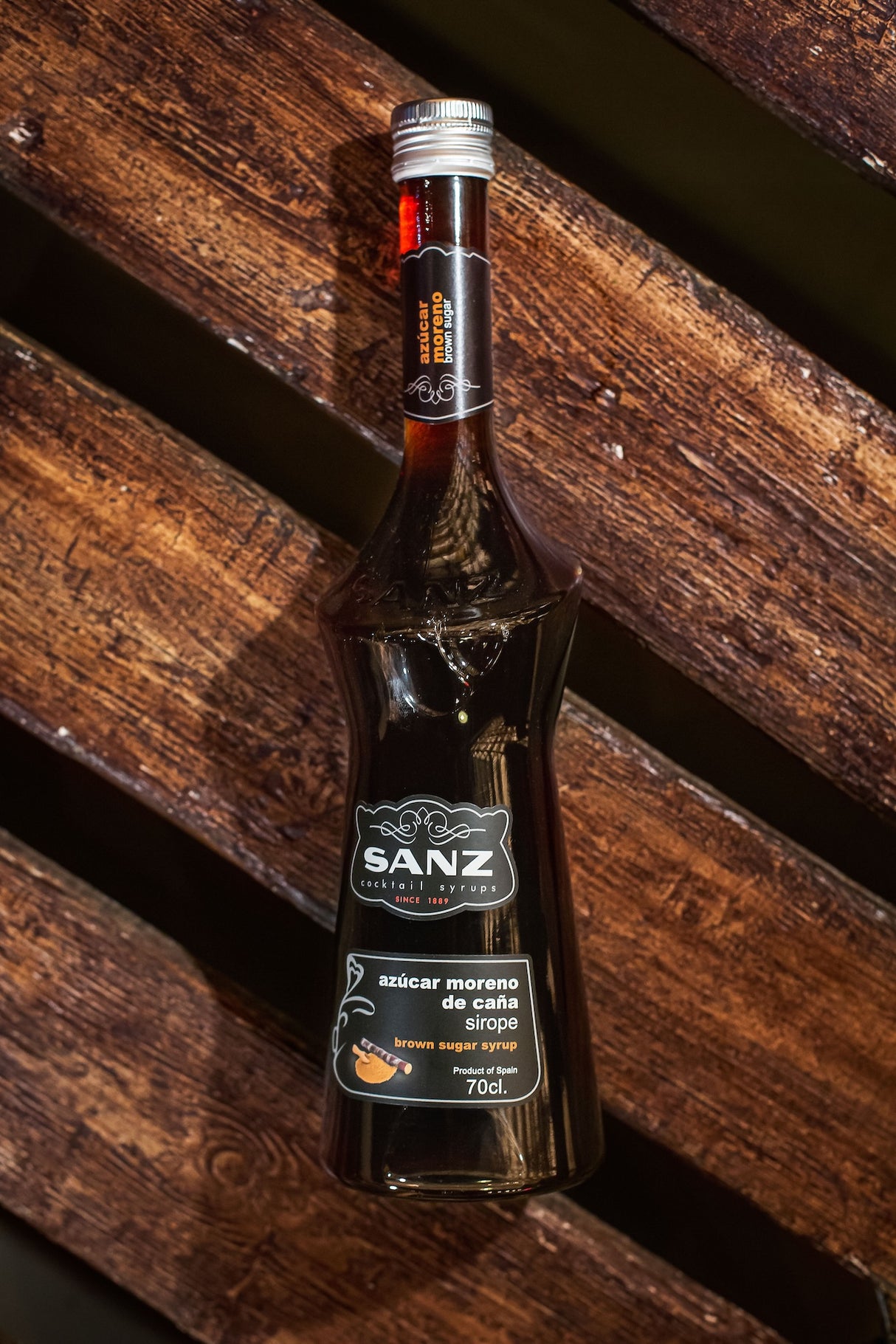 Brauner Rohrzuckersirup Sanz in einer dunklen Flasche auf rustikalem Holzuntergrund, Etikett mit 70 cl-Angabe sichtbar.