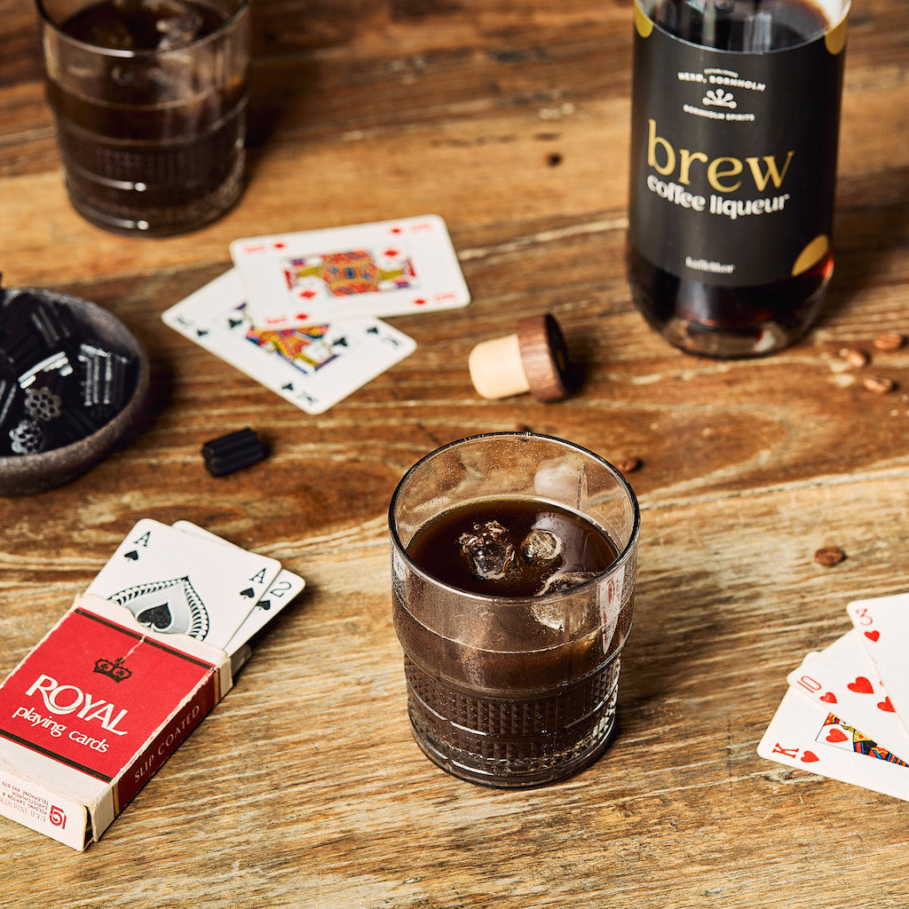 Kaffeelikör mit Eiswürfeln im Tumbler-Glas auf Holztisch, daneben Spielkarten, Flasche und Lakritzstückchen.