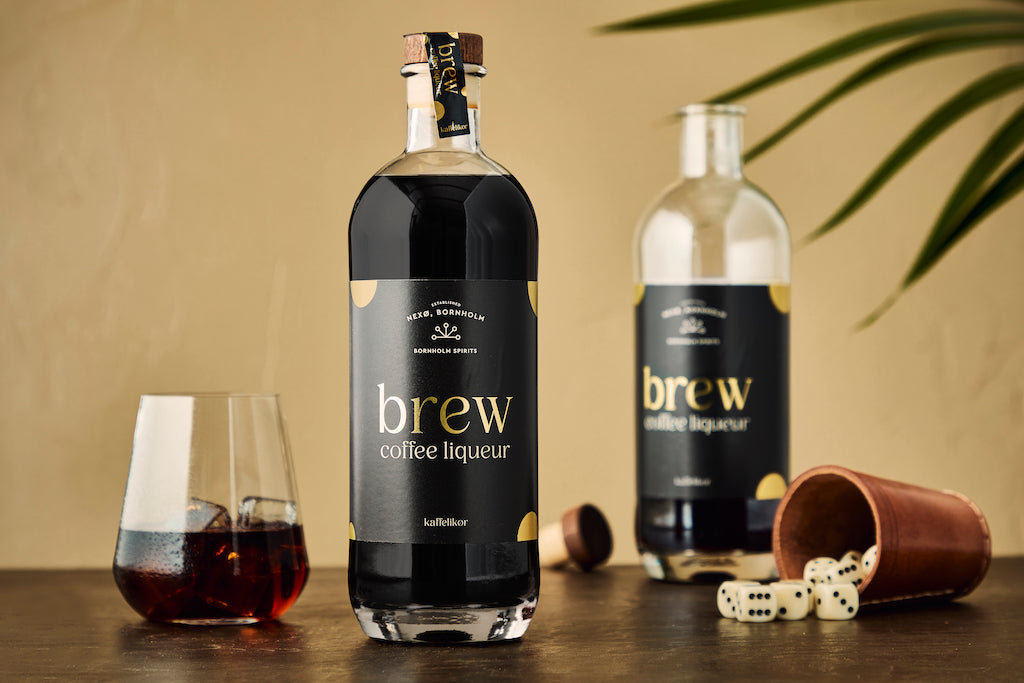 Zwei Flaschen Brew Kaffeelikör mit Glas, Würfelbecher und Spielwürfeln auf dunkler Fläche vor beigem Hintergrund mit Palmenblatt.