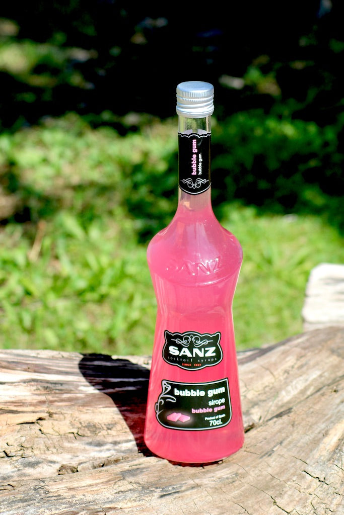 Bubble Gum Sirup Sanz 700 ml in einer pinkfarbenen Flasche mit schwarzem Etikett auf einem Holzstamm.