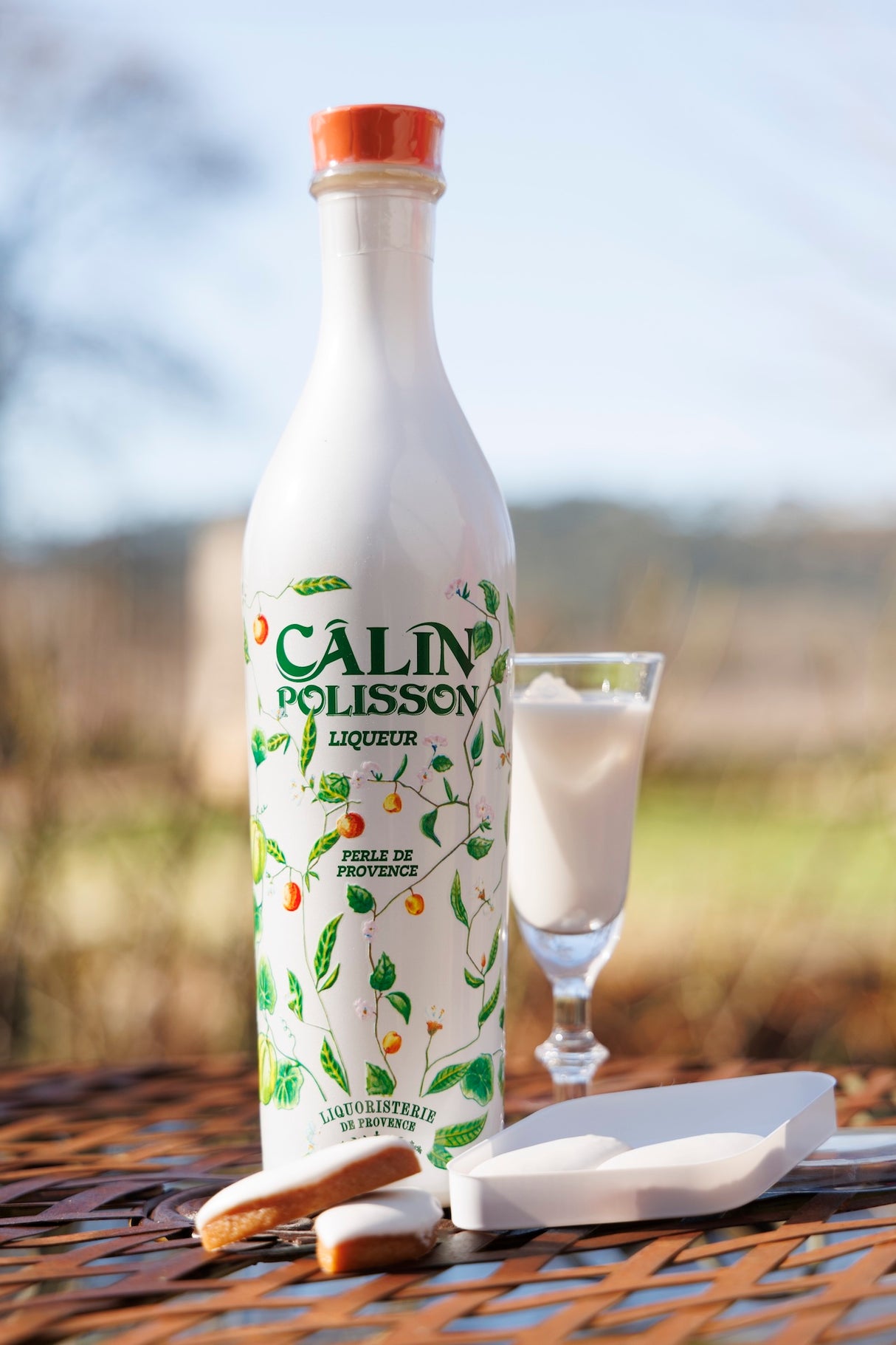 Flasche Calin Polisson Likör mit floraler Gestaltung, daneben Likörglas und Calissons auf Gartentisch vor Naturhintergrund.