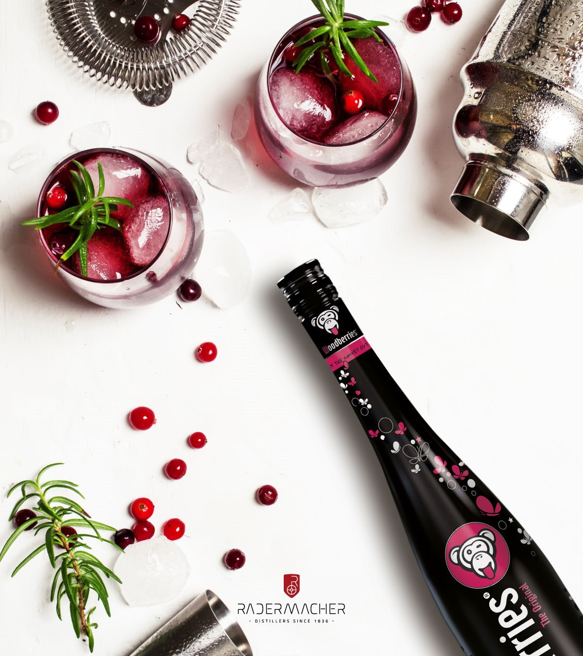 Woodberries-Flasche neben Cocktailgläsern mit Eis, Rosmarin und Beeren, umgeben von Shaker und Barzubehör auf weißem Hintergrund.