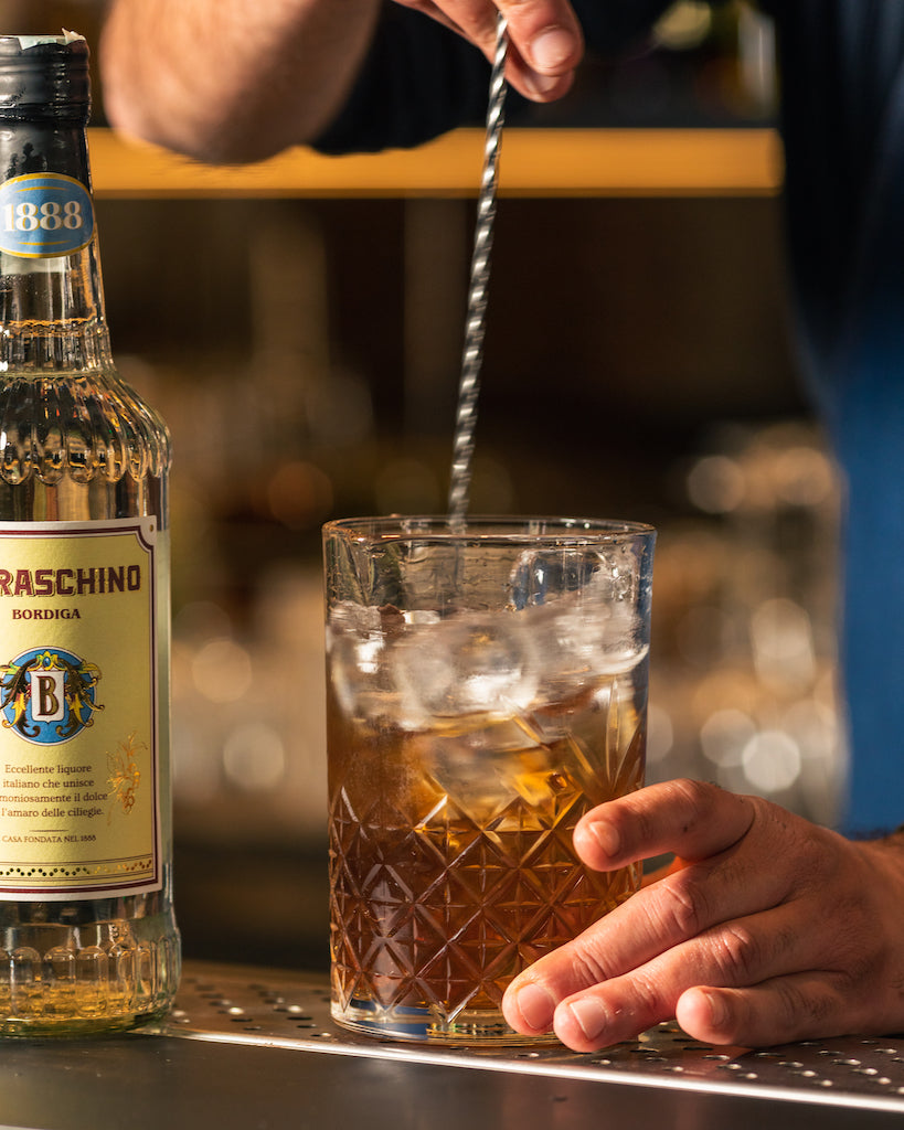 Zubereitung eines Cocktails mit Eis und Rührstab neben einer Flasche Maraschino Bordiga an einer Bar.