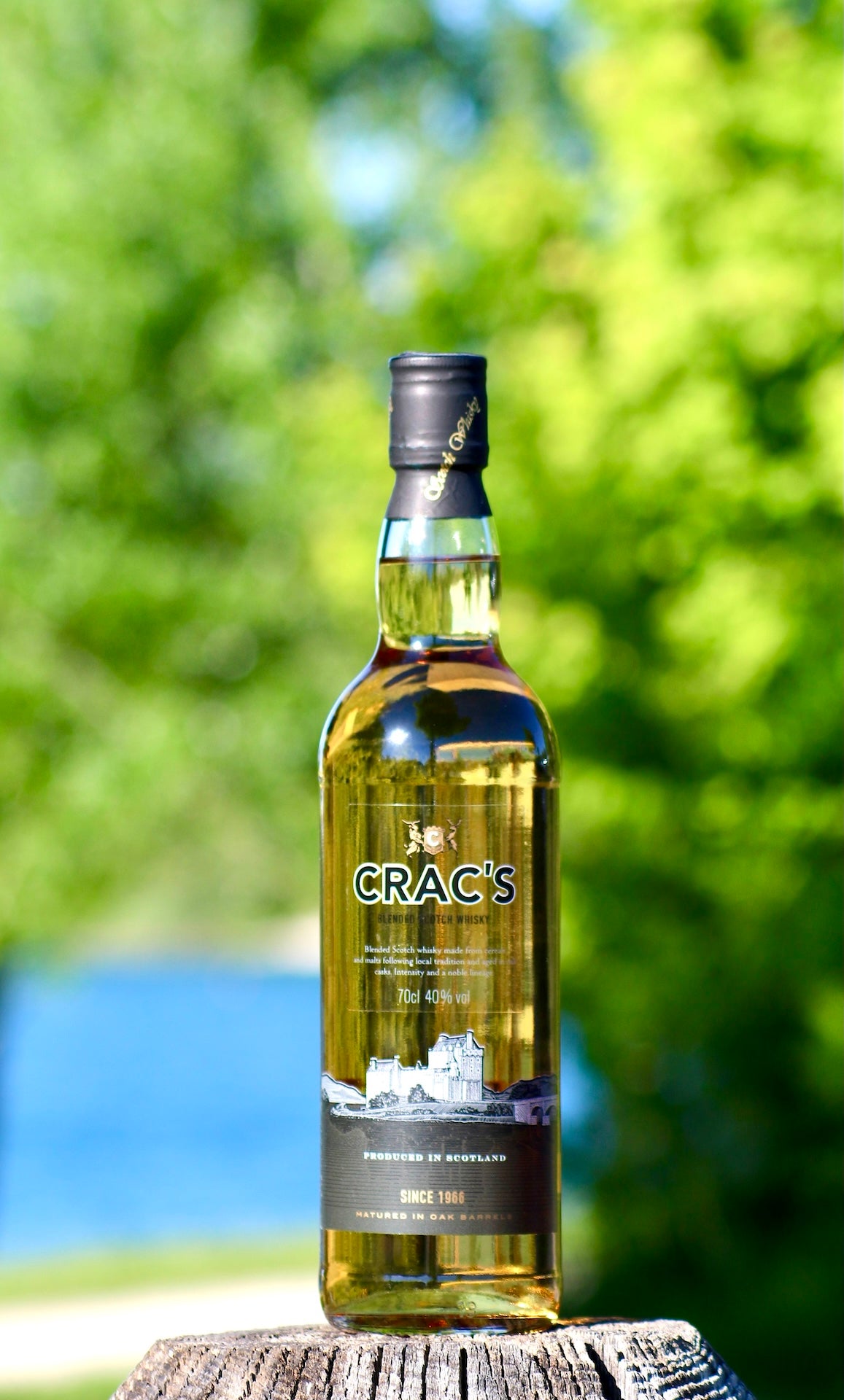 Flasche Crac's Blended Scotch Whisky mit schwarzem Etikett, stehend auf Holz vor grünem Hintergrund und blauem Wasser.