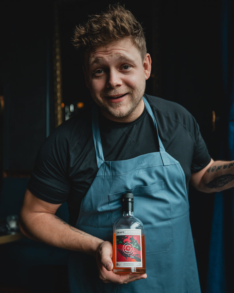 Koch mit blauer Schürze hält eine Flasche Bio-Schnaps Bornholm Rhabarber & Sternanis, dunkler Hintergrund mit gemütlicher Atmosphäre.