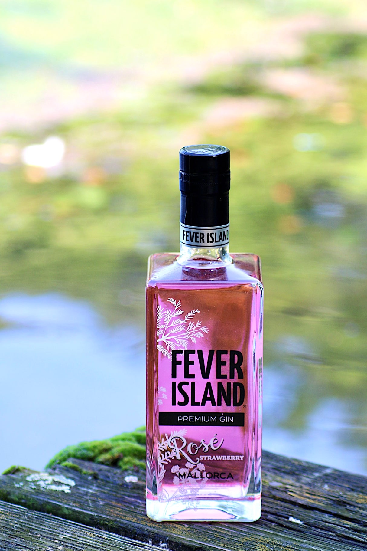 Fever Island Premium Gin Rosé Strawberry in einer rechteckigen Flasche, platziert auf einem hölzernen Untergrund mit unscharfem, natürlichen Hintergrund. Die Flasche zeigt ein elegantes Design mit botanischen Mustern.