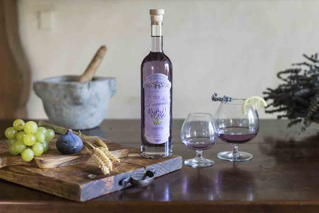 Flasche Fleur de Lavande Likör mit zwei gefüllten Gläsern, daneben Trauben, Feige und Lavendel auf rustikalem Tisch