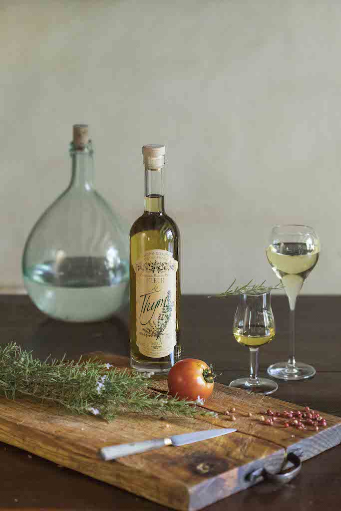 Flasche Fleur de Thym Likör auf Holztisch, Schneidebrett mit Thymian, Tomate und Messer, dazu Gläser mit Likör und Wasserkrug.