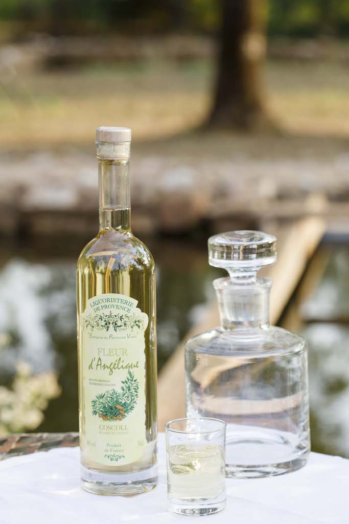 Flasche Fleur d'Angélique Likör neben einer Kristallkaraffe und einem Glas, arrangiert auf weißem Tuch. Im Hintergrund ein Teich.