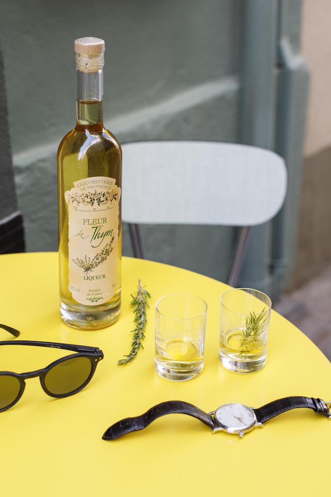 Flasche Fleur de Thym Likör auf gelbem Tisch, zwei Gläser mit Likör und Kräutern, Sonnenbrille, Uhr und Stuhl im Hintergrund.