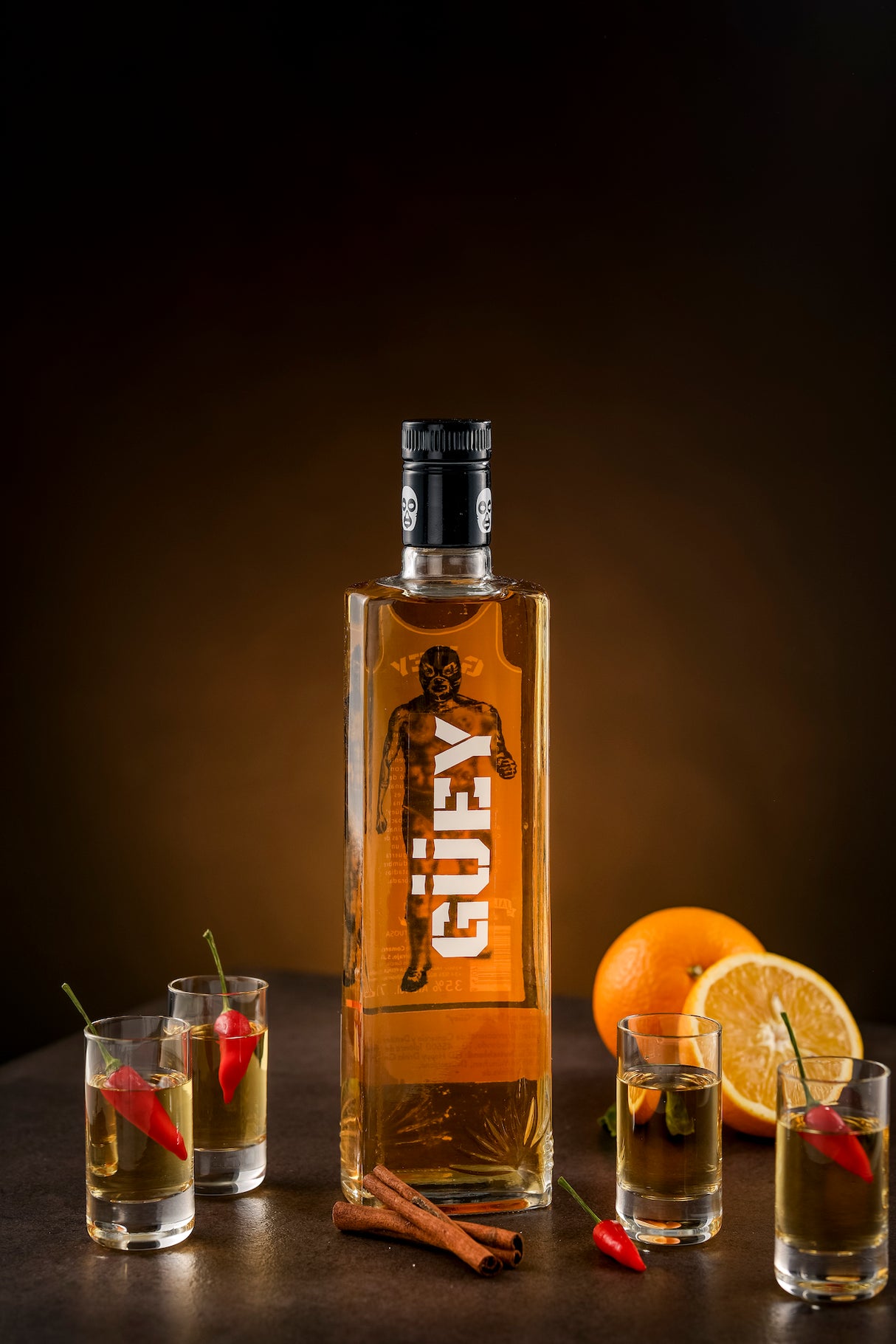 Güey Hot Spicy Gold Tequila, umgeben von Shotgläsern mit Chili, Zimtstangen und frischen Orangenscheiben auf dunklem Hintergrund.