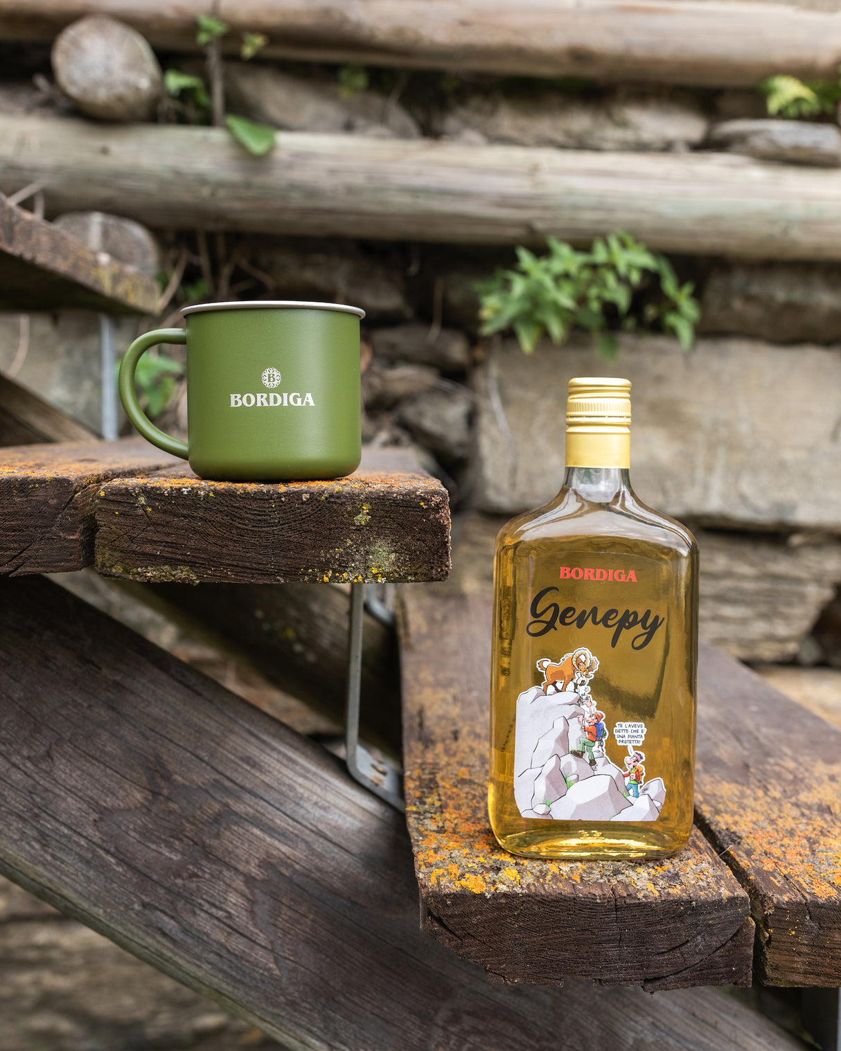 Flasche Bordiga Genepy Likör neben grüner Bordiga-Tasse auf Holzstufen, vor rustikalem Hintergrund mit Steinen und Pflanzen.