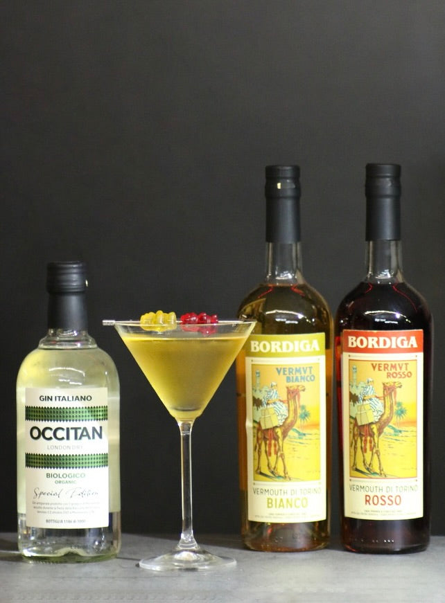 Flaschen von Occitan Gin und Bordiga Vermouth neben einem goldgelben Golden Ermine Cocktail im Martiniglas mit Gummibärchen-Garnitur.