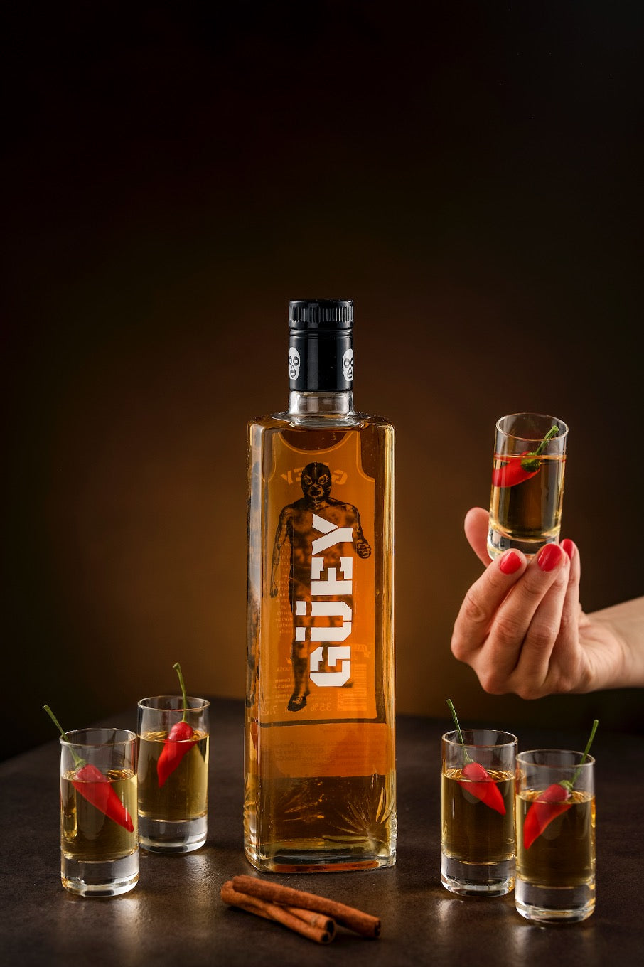 Güey Hot Spicy Gold Tequila, präsentiert mit Chilischoten, Zimtstangen und einem Glas in der Hand vor dunklem Hintergrund.