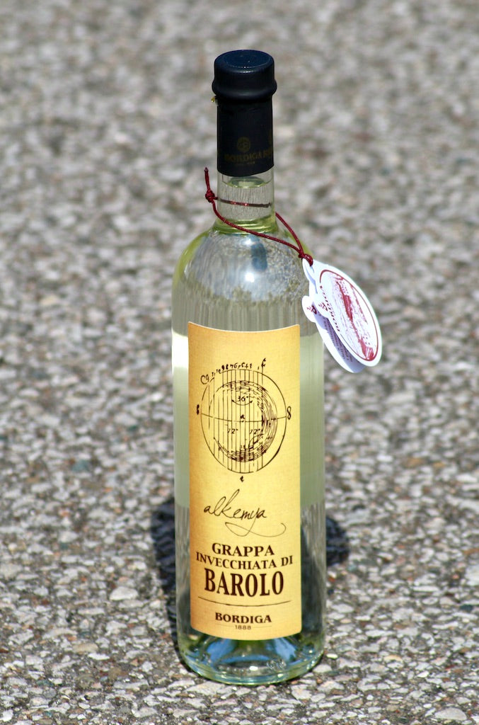 Flasche Grappa Invecchiata di Barolo "Alkemia" von Bordiga, mit Anhänger, präsentiert auf einer gepflasterten Oberfläche.