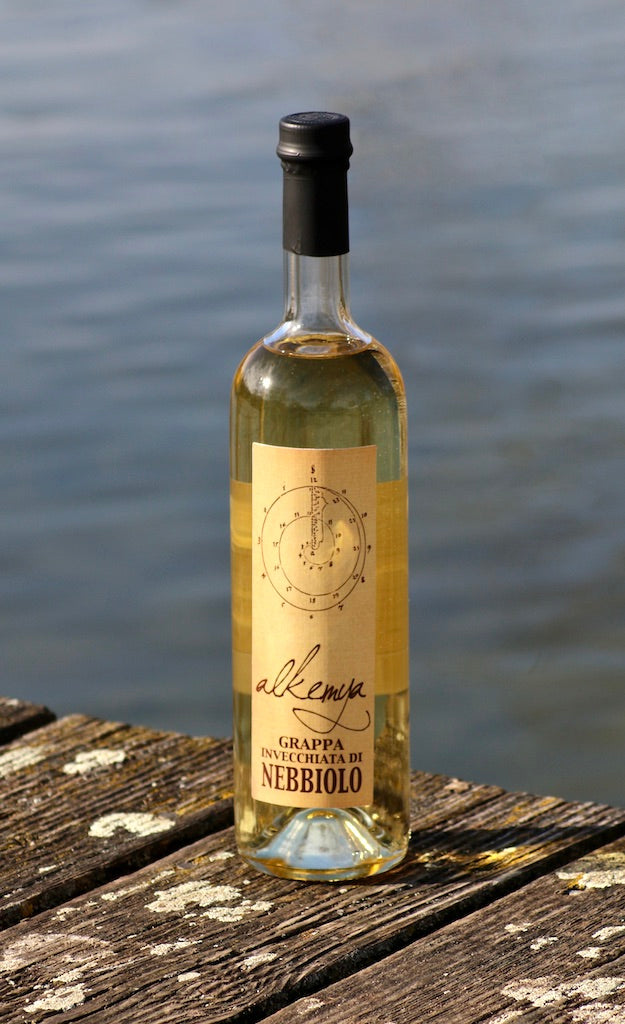 Flasche Grappa Invecchiata di Nebbiolo mit goldfarbenem Etikett, stehend auf einem hölzernen Steg vor einem See.