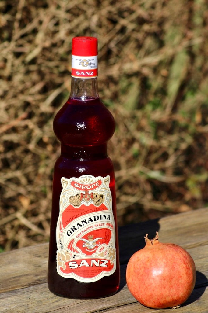 Flasche Sanz Grenadine Sirup 1L und ein Granatapfel auf einer Holzbank, mit unscharfen Sträuchern im Hintergrund.