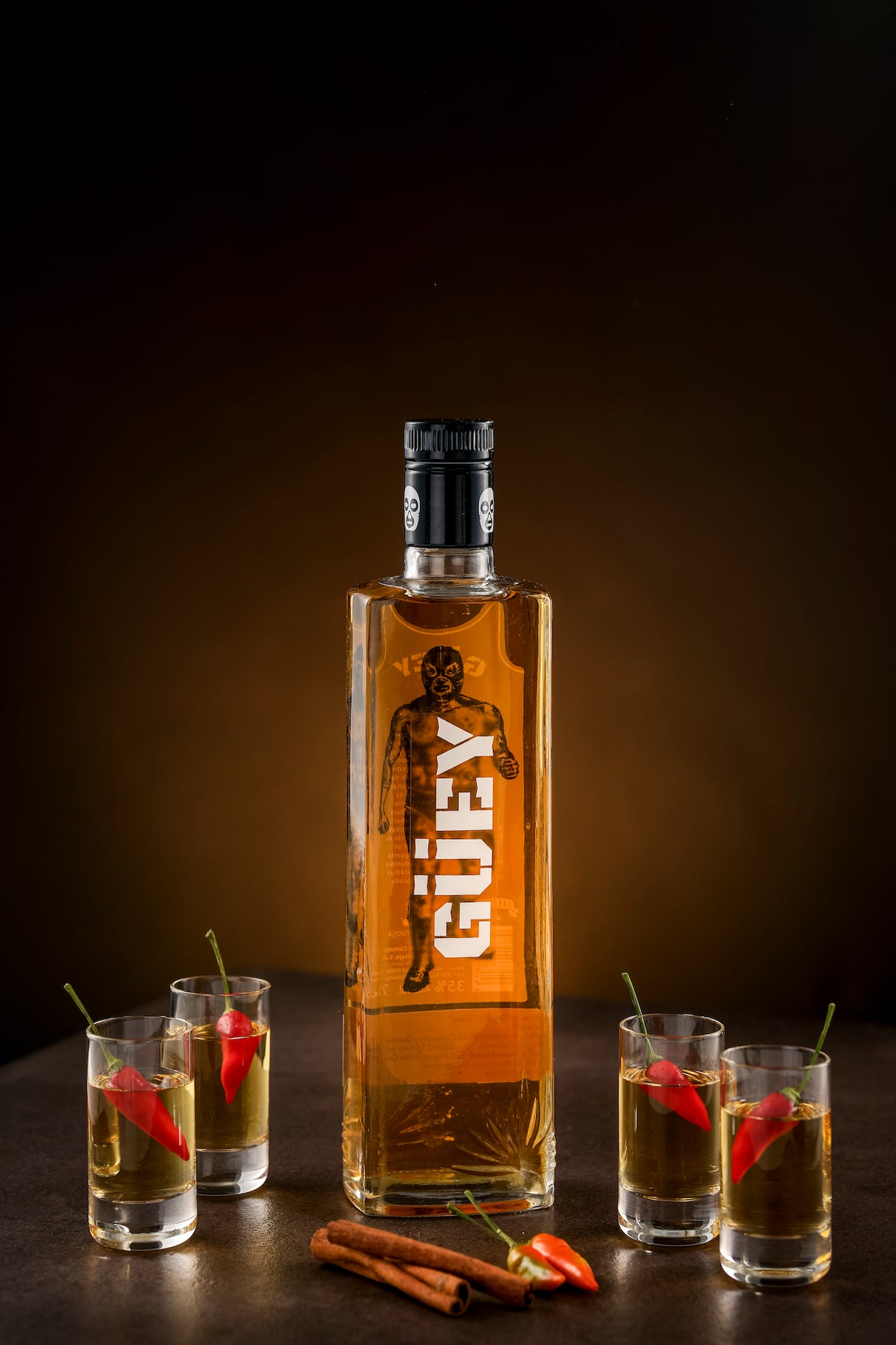 Güey Hot Spicy Gold Tequila, präsentiert mit vier Gläsern, roten Chilischoten, Zimtstangen und einem dunklen, eleganten Hintergrund.