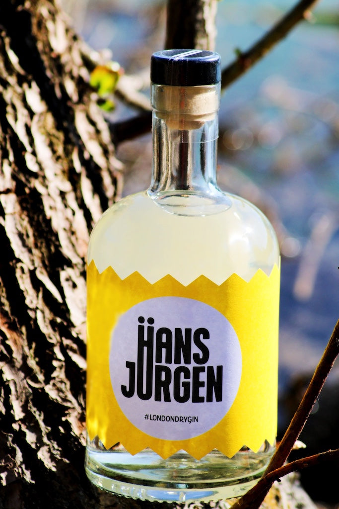 Hans-Jürgen Gin "Heißzeit" Flasche mit gelbem Etikett, fotografiert im Sonnenlicht vor einem Baumstamm, modernes Design.