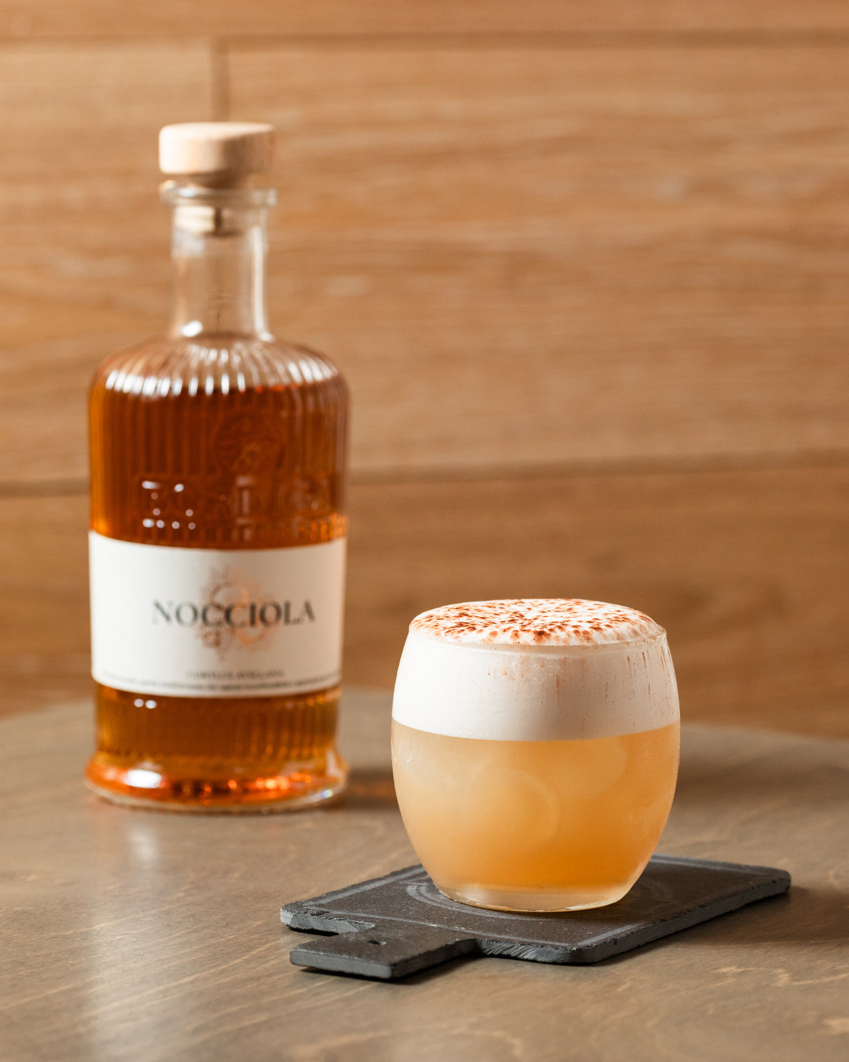 Ein Haselnuss-Sour-Cocktail mit Schaumkrone, serviert auf einer Schieferplatte, im Hintergrund eine Flasche Haselnusslikör.
