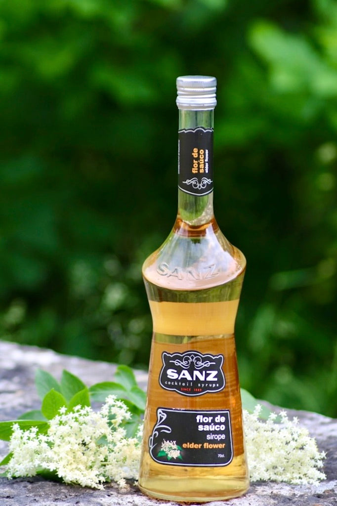 Holunderblütensirup Sanz 0,7L auf Stein mit frischen Holunderblüten, grüne Natur im Hintergrund.