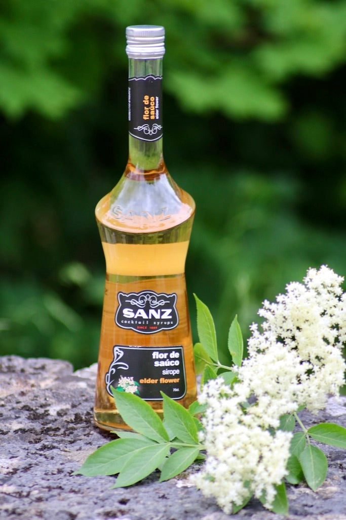 Holunderblütensirup Sanz 0,7L auf Stein mit frischen Holunderblüten, grüner Naturhintergrund.