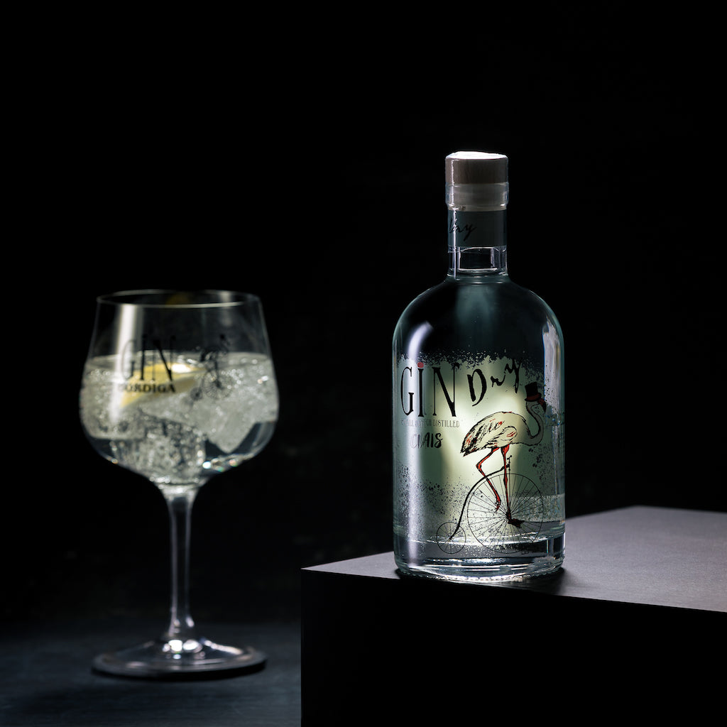 Flasche Bordiga Gin Ciais mit Flamingo-Design auf Podest, daneben ein Gin Tonic im Ballonglas mit Eiswürfeln vor schwarzem Hintergrund.