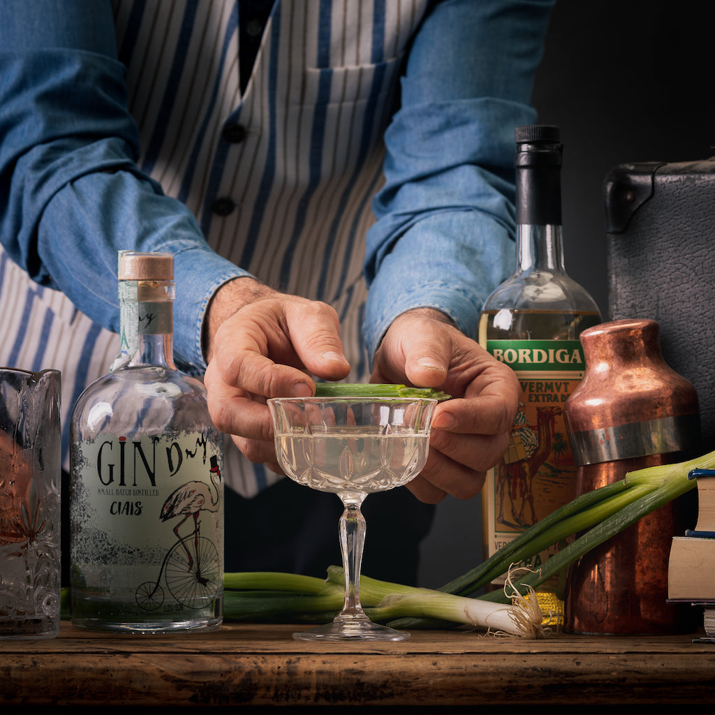 Bartender garniert Cocktailglas mit grünem Blatt, davor Flasche Gin Ciais und Bordiga Vermouth Extra Dry auf rustikalem Tisch.