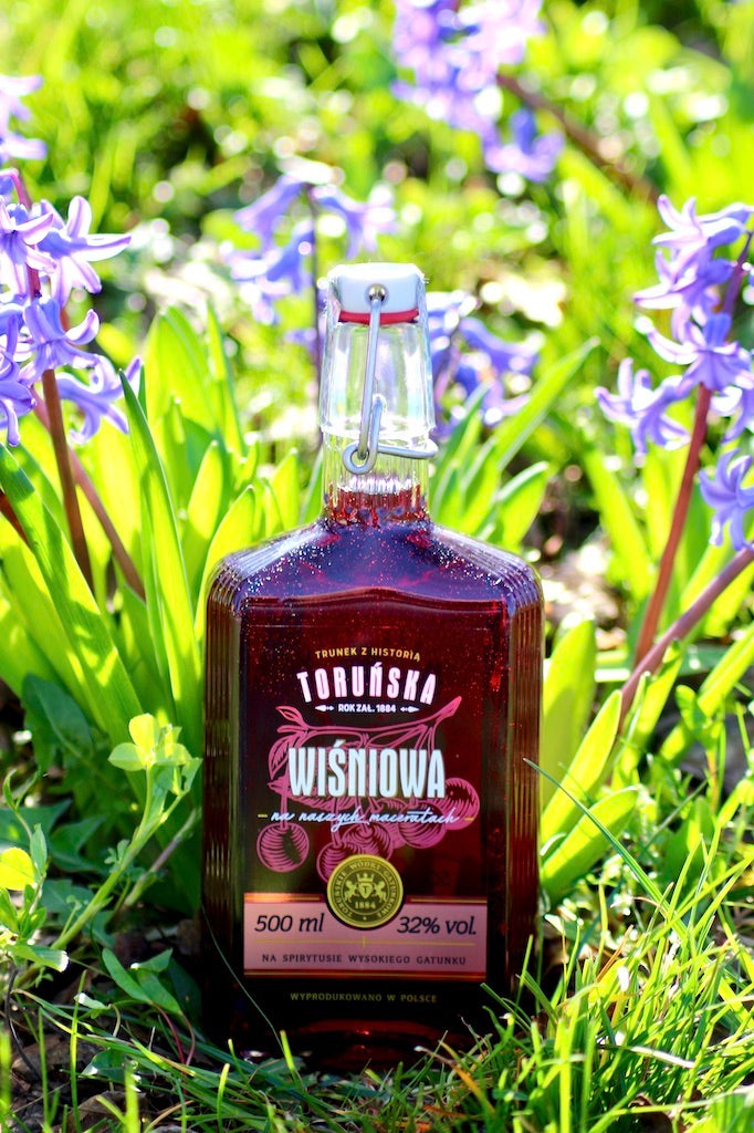 Flasche Toruńska Wiśniowa Kirschlikör, 500 ml, mit Bügelverschluss, inmitten blühender Frühlingsblumen und frischem Grün arrangiert.